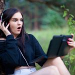 young brunette is sitting in headphones under tree in Park, watching video on tablet computer, she is surprised and scared.